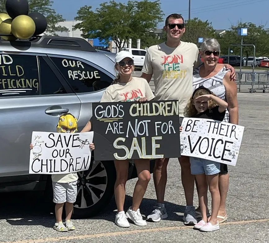 A family holds up signs near their car that say "God's children are not for sale" and "be their voice"