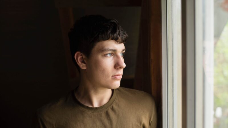 Standing in front of the window and thinking, a young 18-year-old dreams, chooses whether to study or work. Portrait of a charismatic serious guy with blue eyes