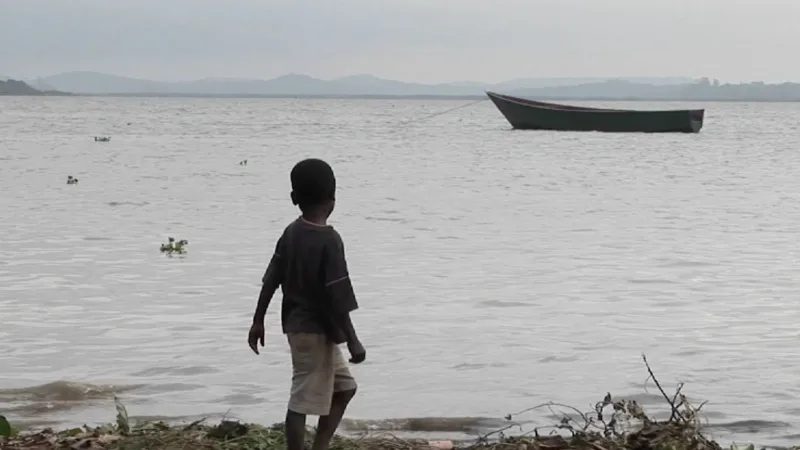 A young boy stands on the shore of a lake with a boat in the background